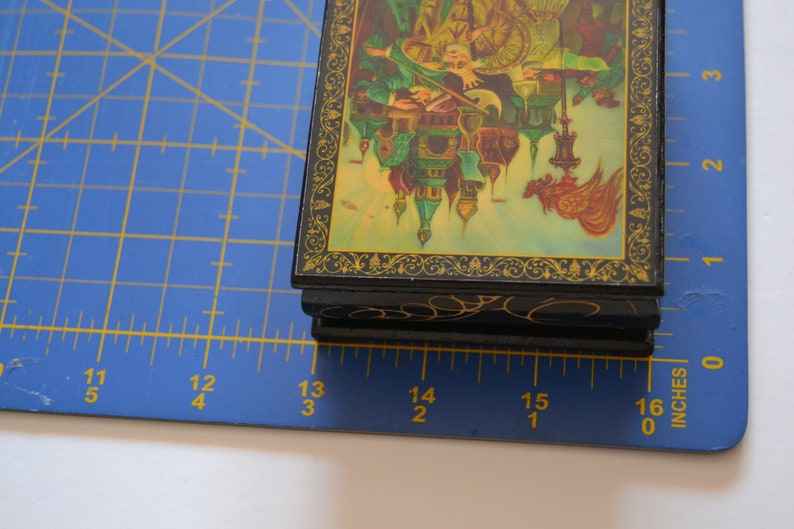 May include: A small, black wooden box with a hinged lid. The lid is decorated with a colorful painting of a scene with buildings, people, and a bird. The box is about 4 inches wide and 3 inches tall.