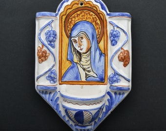 May include: A white ceramic wall plaque with a blue and yellow image of a woman in a blue robe. The plaque has a small shelf at the bottom and is decorated with grape vines on the sides.