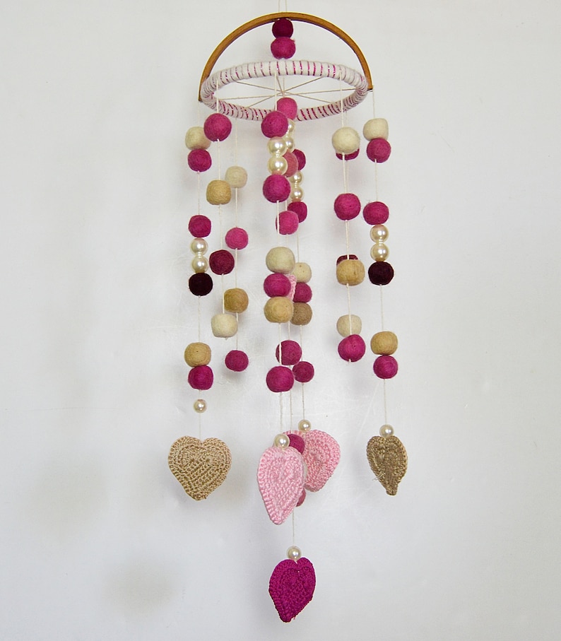 May include: A pink and cream felt ball mobile with crocheted hearts hanging from a wooden hoop. The mobile is decorated with pearl beads.