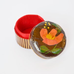 Puede incluir: Una pequeña caja de madera con un interior rojo y un diseño floral pintado en la tapa. La flor es naranja y amarilla con hojas verdes.