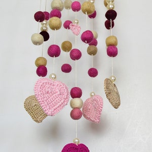 May include: A pink and beige felt ball mobile with crocheted hearts hanging from it. The mobile is made of felt balls, crocheted hearts, and pearl beads.