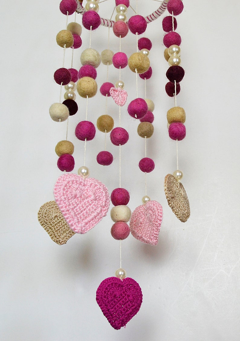May include: A pink and beige felt ball mobile with crocheted hearts hanging from it. The mobile is made of felt balls, crocheted hearts, and pearl beads.