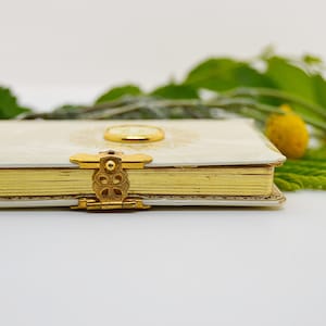 May include: A white journal with a gold clasp and a gold decorative design on the cover. The journal is closed and lying on a white surface. There are green leaves and yellow flowers in the background.