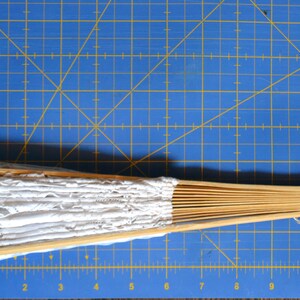 May include: A white lace fan with a wooden handle. The fan is closed and has a white tassel.