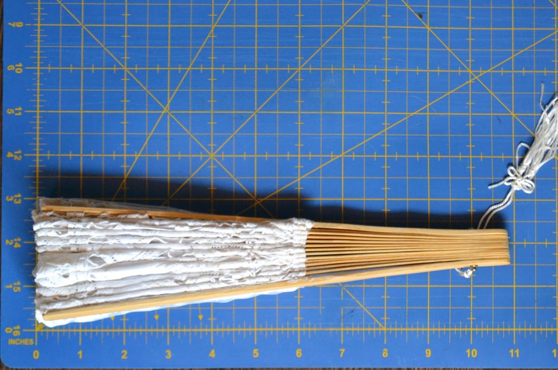 May include: A white lace fan with a wooden handle. The fan is closed and has a white tassel.
