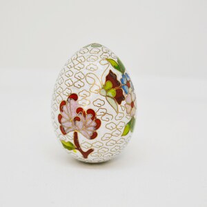 May include: A white egg-shaped decorative object with a floral design. The egg is decorated with red, pink, blue, green, and gold enamel. The background is a white, textured pattern.