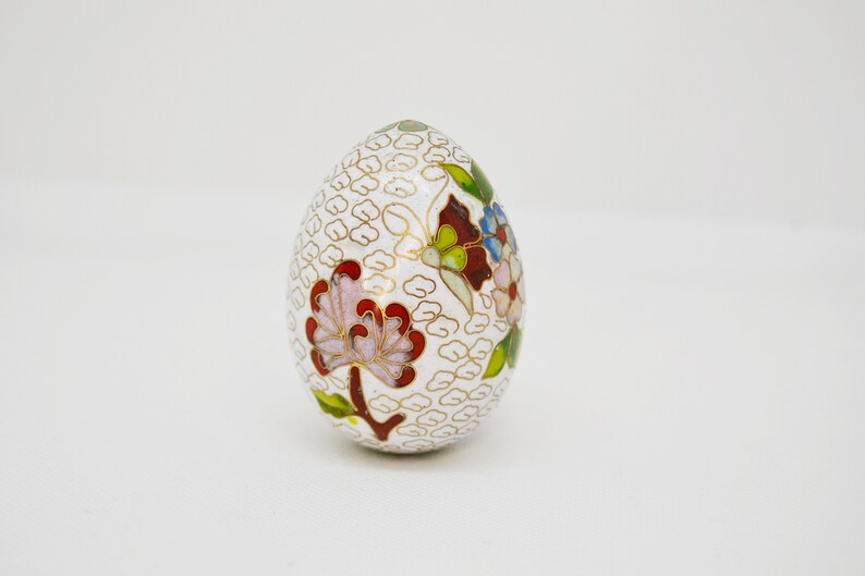 May include: A white egg-shaped decorative object with a floral design. The egg is decorated with red, pink, blue, green, and gold enamel. The background is a white, textured pattern.