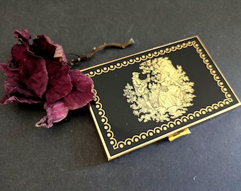 May include: A black compact with a gold border and a gold illustration of a couple in a garden. A dried burgundy flower and stem are next to the compact on a dark surface.