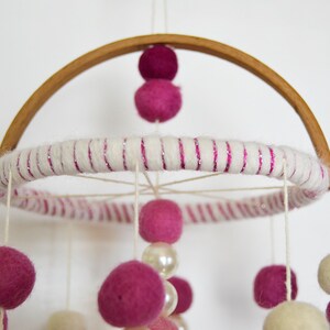 May include: A pink and white baby mobile with felt balls and wooden beads hanging from a wooden hoop. The hoop is wrapped in white yarn with pink glitter.