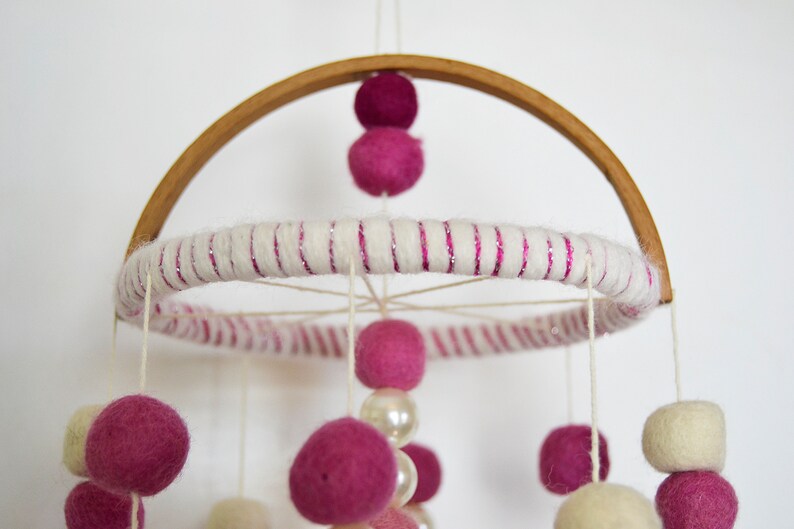 May include: A pink and white baby mobile with felt balls and wooden beads hanging from a wooden hoop. The hoop is wrapped in white yarn with pink glitter.