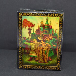 May include: A black lacquered wooden box with a colorful painted scene of a city with a rooster on top of a lamppost. The scene is framed with a gold border.