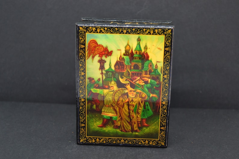 May include: A black lacquered wooden box with a colorful painted scene of a city with a rooster on top of a lamppost. The scene is framed with a gold border.