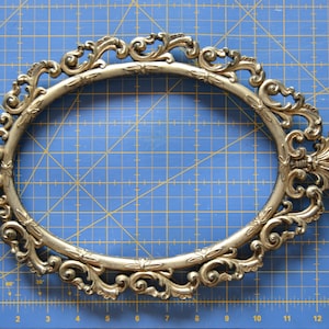 May include: A gold-colored, ornate oval picture frame with a decorative, scroll-like design. The frame is made of metal and has a smooth, polished finish.