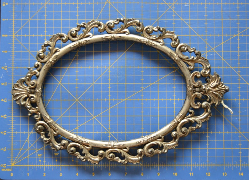 May include: A gold-colored, ornate oval picture frame with a decorative, scroll-like design. The frame is made of metal and has a smooth, polished finish.