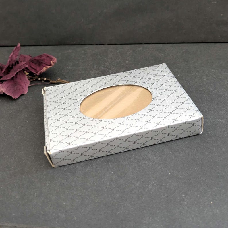 Puede incluir: Una caja rectangular plateada con un dise&ntilde;o estampado y una ventana ovalada que revela un art&iacute;culo de color claro. La caja est&aacute; cerrada y descansa sobre una superficie oscura. Una flor morada seca est&aacute; en el fondo.