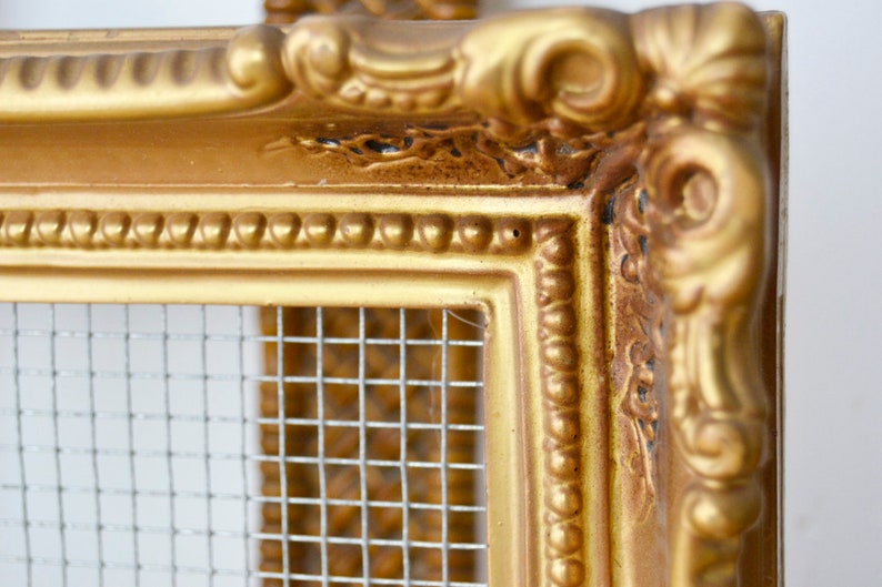 May include: An ornate picture frame in a gold colour with a wire mesh insert. The frame has a decorative, ornate design.