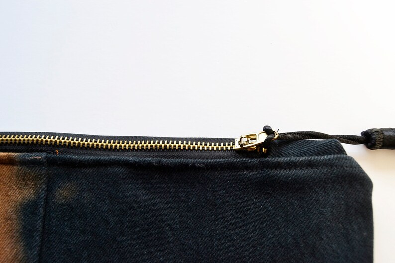 May include: A black canvas zippered pouch with a gold zipper and a black drawstring. The pouch has a brown and black tie-dye pattern on one side.