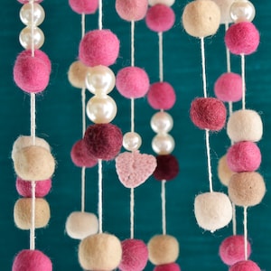 May include: A pink, white, and beige felt ball mobile with a crocheted heart hanging from the center. The mobile is suspended from a white string.