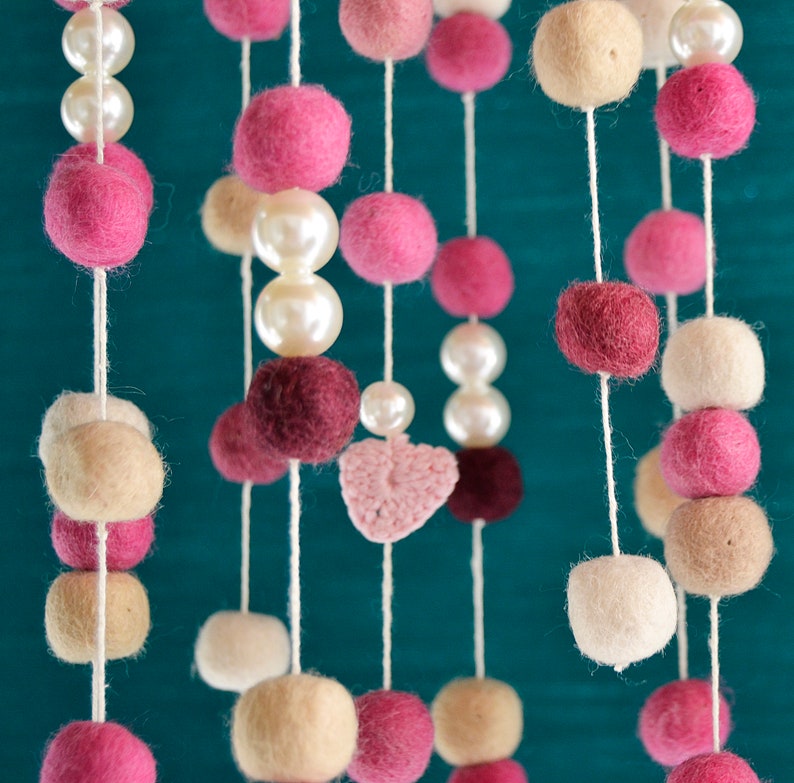 May include: A pink, white, and beige felt ball mobile with a crocheted heart hanging from the center. The mobile is suspended from a white string.