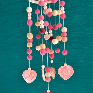 May include: A pink and beige baby mobile with crocheted hearts and felt balls hanging from a wooden ring.
