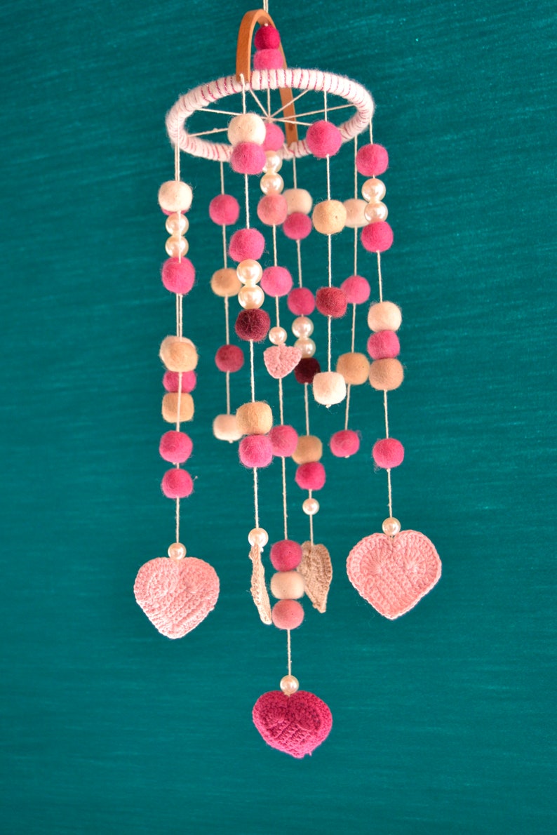 May include: A pink and beige baby mobile with crocheted hearts and felt balls hanging from a wooden ring.