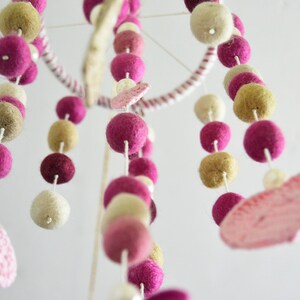 May include: A pink and beige felt ball mobile with two pink felt hearts hanging from it. The mobile is made of felt balls and is strung on white string.
