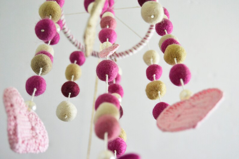 May include: A pink and beige felt ball mobile with two pink felt hearts hanging from it. The mobile is made of felt balls and is strung on white string.