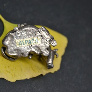 May include: A silver alpaca figurine with a textured surface, sitting on a yellow leaf. The figurine has the word "ALPACA" written on its side.