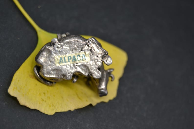 May include: A silver alpaca figurine with a textured surface, sitting on a yellow leaf. The figurine has the word "ALPACA" written on its side.
