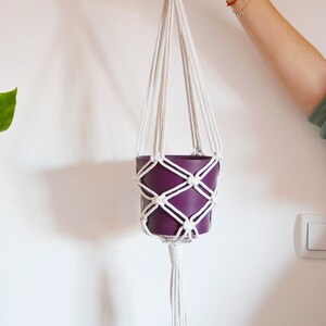 Puede incluir: Un macetero de macramé blanco con una maceta morada colgando.