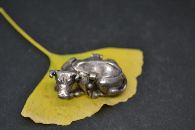 May include: A silver cow figurine with a detailed design, resting on a yellow ginkgo leaf.