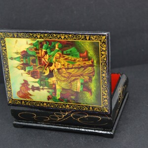 May include: A black wooden box with a hinged lid. The lid is decorated with a colorful painting of a scene from a Russian fairy tale. The box is about 4 inches tall and 3 inches wide.