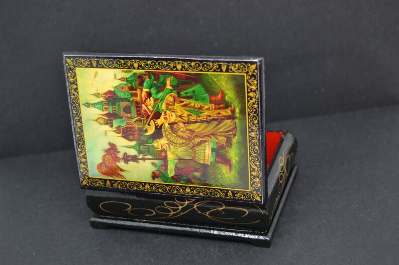 May include: A black wooden box with a hinged lid. The lid is decorated with a colorful painting of a scene from a Russian fairy tale. The box is about 4 inches tall and 3 inches wide.