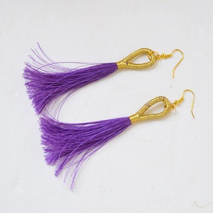 May include: A pair of long, purple tassel earrings with gold-colored metal accents. The earrings feature a loop design at the top and a hook closure. The tassels are made of thin, purple threads.