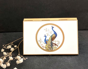 May include: A rectangular box with a gold-colored frame and a white front panel. The panel features a circular design with two blue and gold peacocks perched on a branch. The box is on a dark surface.
