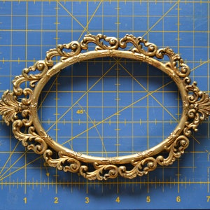 May include: A gold-colored ornate oval picture frame with a detailed, intricate design. The frame is on a blue gridded surface.