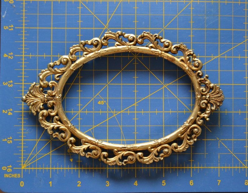 May include: A gold-colored ornate oval picture frame with a detailed, intricate design. The frame is on a blue gridded surface.