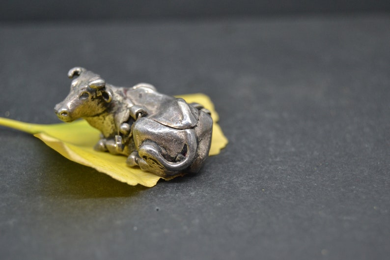May include: A silver figurine of a cow lying down on a yellow leaf. The cow is detailed with intricate carvings and has a realistic appearance.