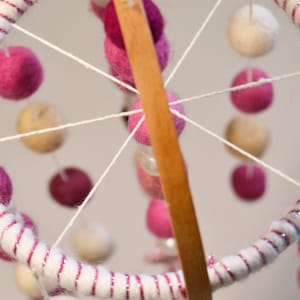 May include: A white and pink baby mobile with felt balls hanging from a wooden dowel. The balls are arranged in a circular pattern and are connected by white string.
