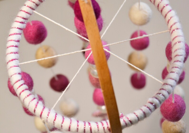 May include: A white and pink baby mobile with felt balls hanging from a wooden dowel. The balls are arranged in a circular pattern and are connected by white string.