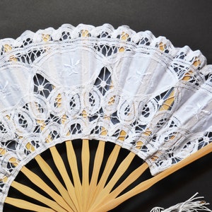 May include: A white lace fan with a wooden handle. The lace is delicate and intricate, with a floral pattern. The fan is closed, but the wooden ribs are visible.