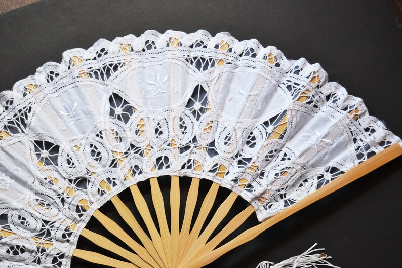 May include: A white lace fan with a wooden handle. The lace is delicate and intricate, with a floral pattern. The fan is closed, but the wooden ribs are visible.