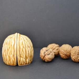Puede incluir: Un cascanueces de cerámica con forma de nuez, en tonos marrones, dividido en dos mitades, junto a varias nueces enteras. El cascanueces tiene un acabado brillante y una textura detallada.