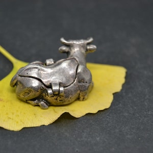 May include: A silver cow figurine with a hinged lid, resting on a yellow ginkgo leaf. The cow is detailed with a textured surface and has a small opening on its back.