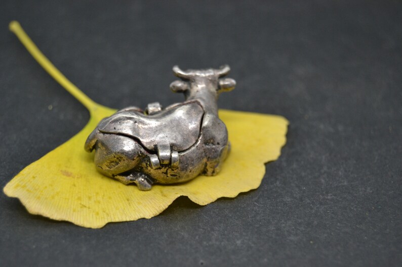 May include: A silver cow figurine with a hinged lid, resting on a yellow ginkgo leaf. The cow is detailed with a textured surface and has a small opening on its back.