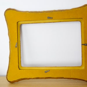 May include: A yellow picture frame with a decorative, ornate border. The frame has four metal hooks on the back.