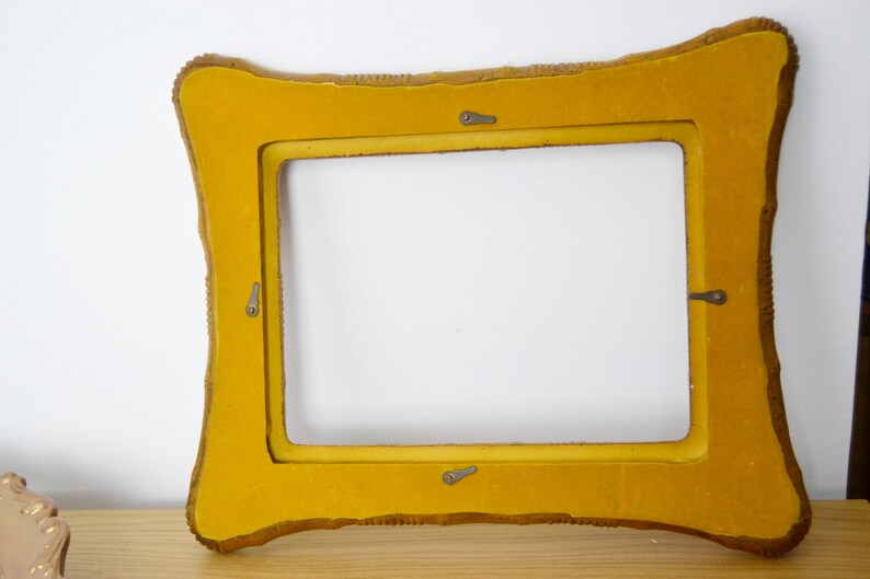 May include: A yellow picture frame with a decorative, ornate border. The frame has four metal hooks on the back.