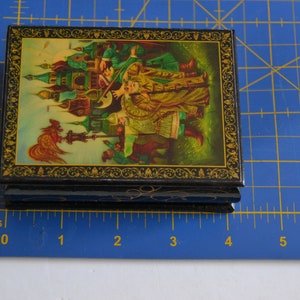 May include: A small, black wooden box with a hinged lid. The lid is decorated with a colorful painting of a Russian folk scene. The box is about 4 inches wide and 3 inches tall.