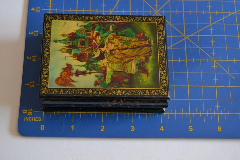 May include: A small, black wooden box with a hinged lid. The lid is decorated with a colorful painting of a Russian folk scene. The box is about 4 inches wide and 3 inches tall.