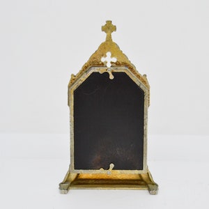 May include: Ornate gold-colored metal picture frame with a cross at the top. The frame has a dark, rectangular insert and a decorative, architectural design. The frame is standing upright on a white surface.
